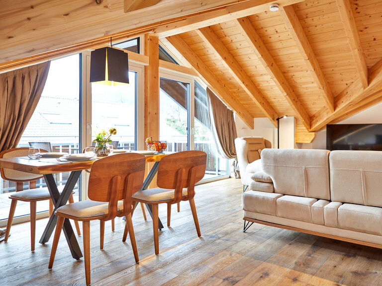 Ein eleganter Holzesstisch mit komfortablen Sitzgelegenheiten und eine gemütliche Couch mit Fernseher vor einer großen Fensterfront.
